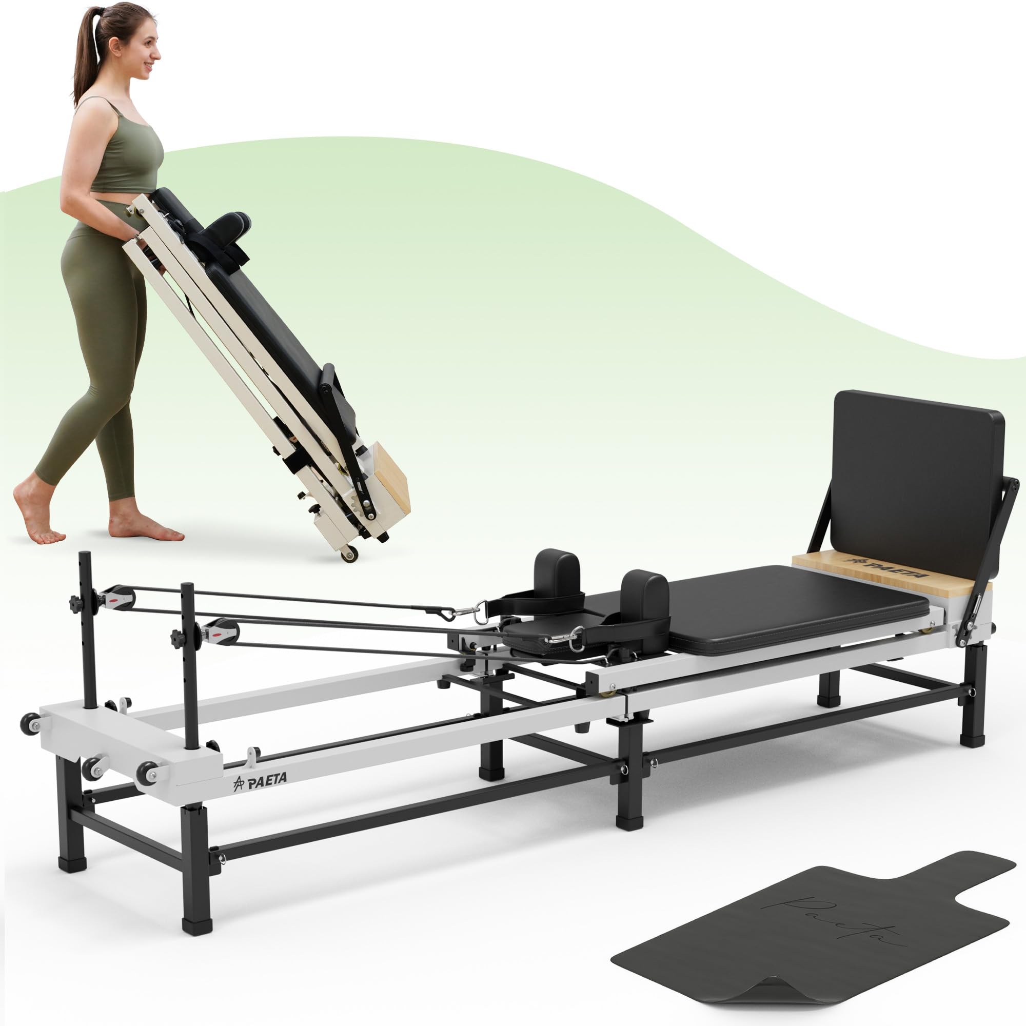 A woman in workout clothes is folding and carrying a compact Pilates reformer. Another reformer is set up in front of her with a black mat placed nearby on the floor.