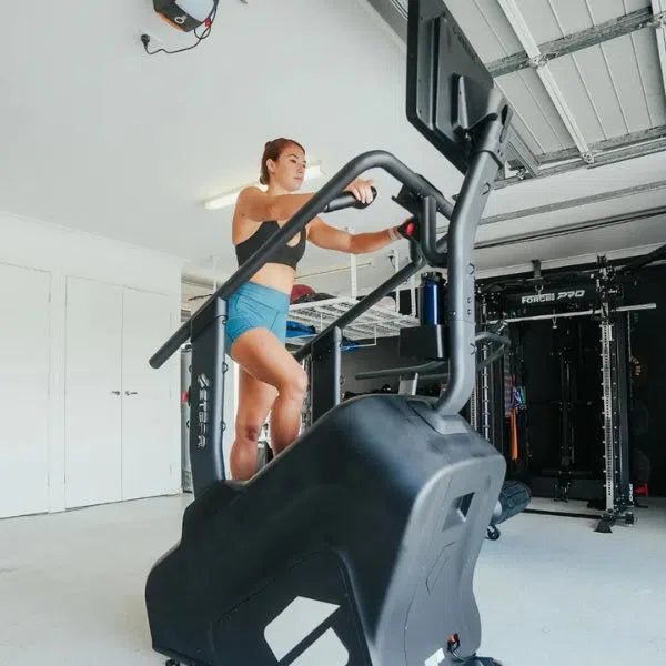 A woman exercises on the STEPR PRO+ Commercial Stair Climber by STEPR in her home gym, wearing a black sports bra and teal shorts. Focused, she interacts with the HD touchscreen amid fitness machines, set against gray floors and white walls.