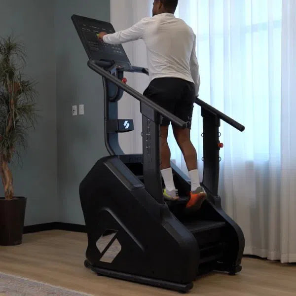 A person wearing a white shirt and black shorts is intensely training on the STEPR PRO Classic Stair Climber by STEPR. The room, with light walls, a big window with sheer curtains, and a potted plant, provides an invigorating workout setting.