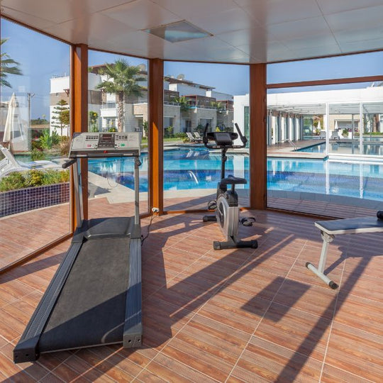 Modern indoor gym with large windows showing an outdoor pool. Features a treadmill, stationary bike, and bench on a tiled floor. Bright natural lighting enhances the serene and inviting atmosphere.