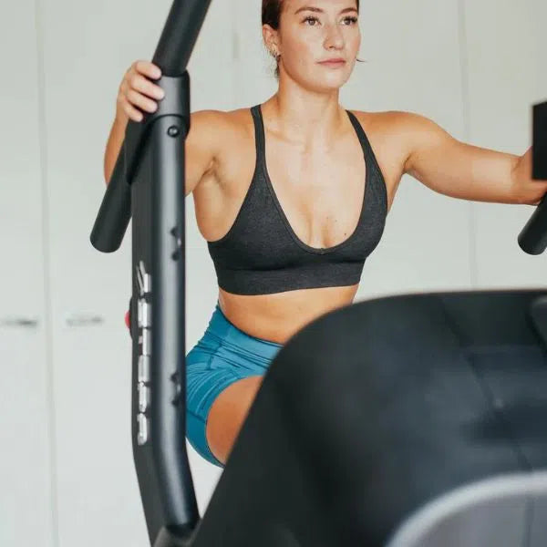 A woman in workout attire on the STEPR PRO+ Commercial Stair Climber with an HD touchscreen wears a black sports bra and blue shorts. Indoors, the blurred background highlights her determined expression, showcasing the intensity of her workout.