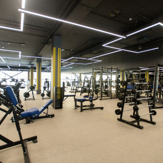 A spacious gym with various weightlifting equipment, including benches, racks, and dumbbells. The ceiling features modern, geometric lighting, and the floor is covered with a light gray carpet. Walls have bold, vertical yellow stripes.