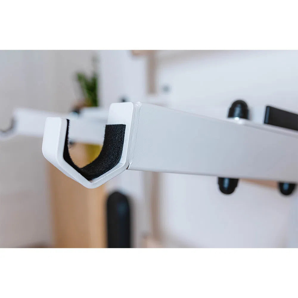 Close-up of a BenchK Home Wall Bars w/ Flex Pull-Up Bar (731) showing a metal hook mounted on a wall. It meets European safety standards and has a U-shaped design with padded inner surfaces. The blurred background subtly hints at plant and wooden elements.