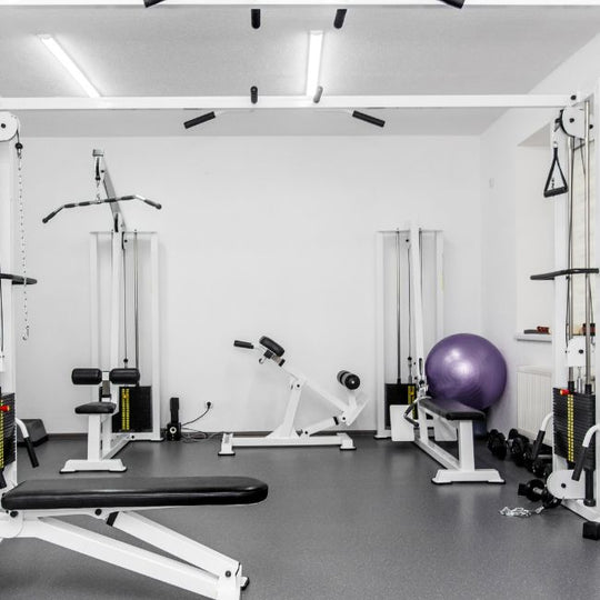 A compact gym room featuring various workout equipment, including a bench press, leg extension machine, exercise ball, and weight stack machines. The walls are white, and the floor is gray with lighting from the ceiling.
