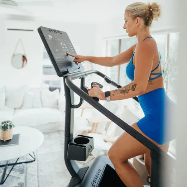 In her bright home gym with white walls, a woman in blue uses the STEPR Classic Stair Climber. The space includes a round table, light sofa, and large windows. Focused on cardio, she adjusts the STEPR screen settings.