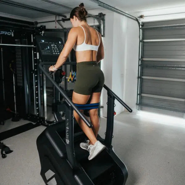 A woman in a white sports bra and green shorts works out on a STEPR Classic Stair Climber in her home gym. The space, featuring a gray garage door and diverse fitness gear, forms the ideal setting for staying fit.