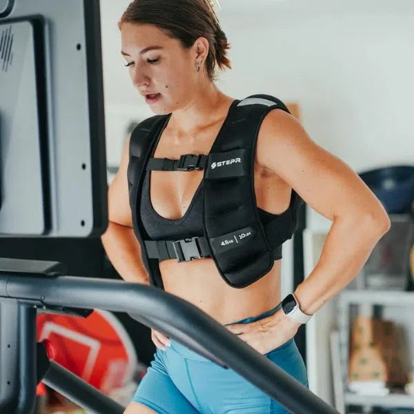 A woman exercises on the STEPR PRO+ Commercial Stair Climber, wearing a black weighted vest. Focused and determined, her hair tied back, she sports a black tank top, blue shorts, and a smartwatch. An HD touchscreen adorns the fitness machine in front of her.