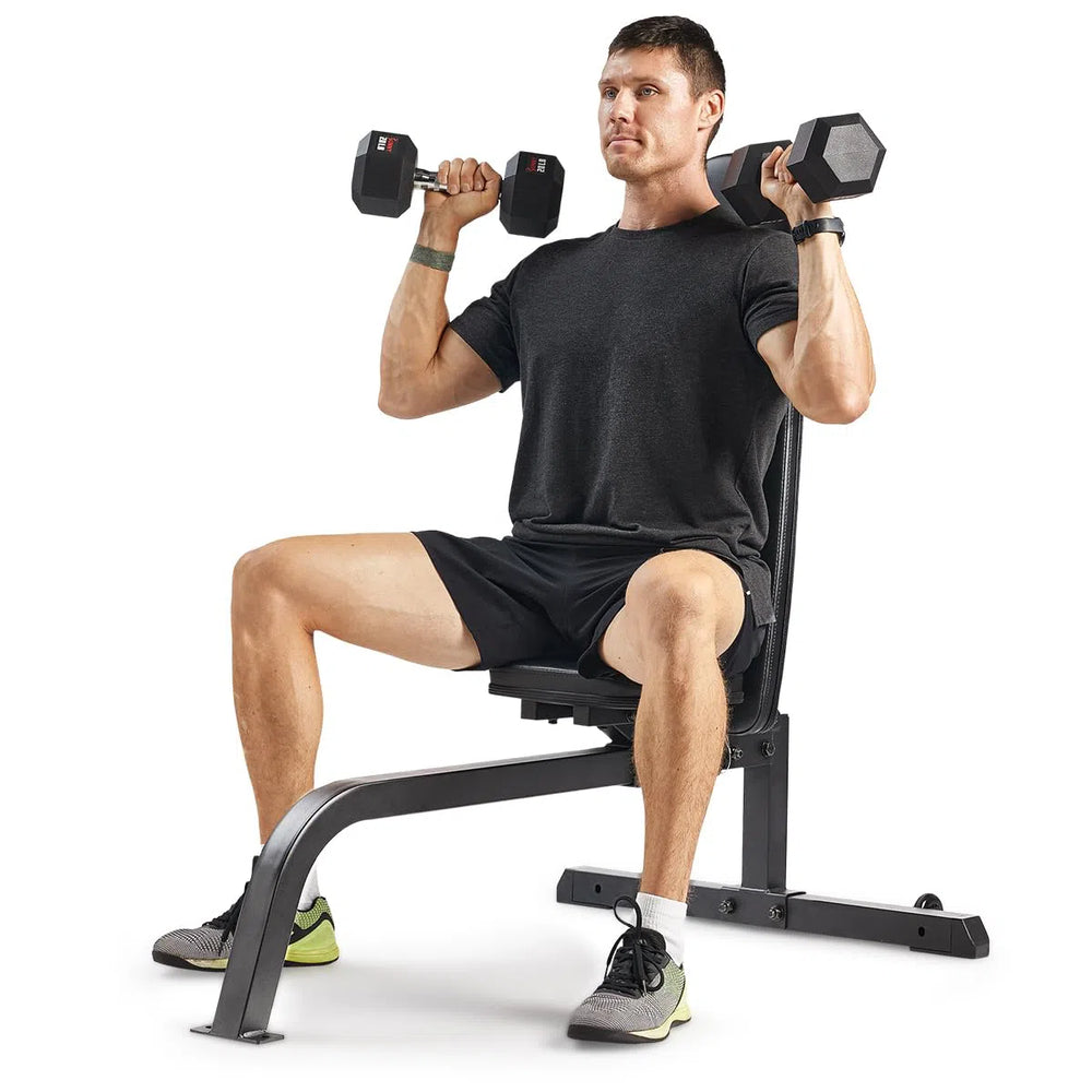 A man uses the Sunny Health & Fitness Incline Bench for a shoulder press with dumbbells, wearing a black t-shirt, black shorts, and green-gray sneakers against a plain white background.