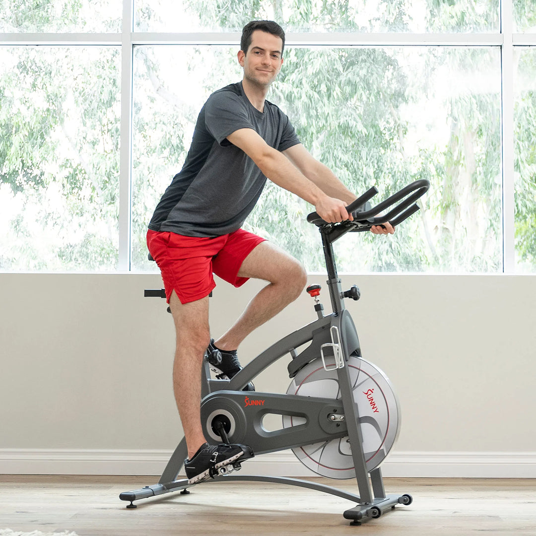 A person in gray shirt and red shorts is invigorated on the Sunny Health & Fitness Synergy Series Magnetic Indoor Cycling Exercise Bike. Positioned on wooden flooring, the large windows reveal green trees outside, merging natures tranquility into the workout.