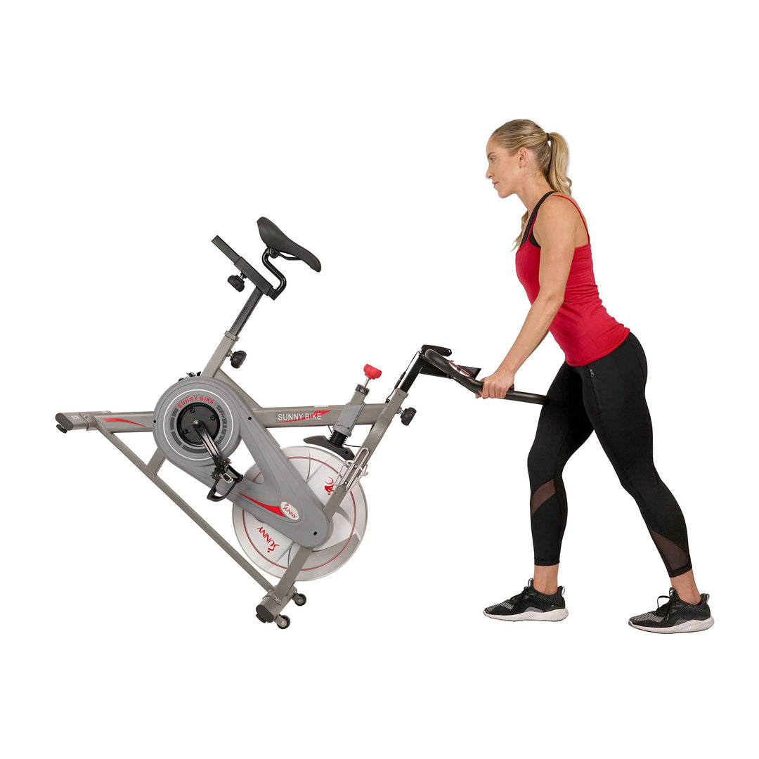 A woman in a red tank top and black leggings is maneuvering the Sunny Health & Fitness Synergy Series Magnetic Indoor Cycling Exercise Bike, actively gripping the handlebars and tilting it on its wheels with purpose.