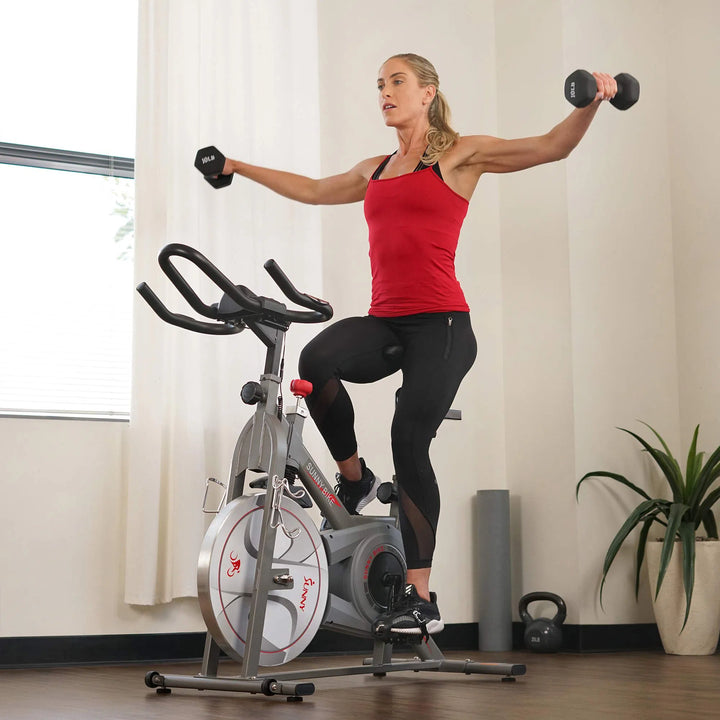 In a sunlit room with a potted plant, a woman in a red tank top and black leggings uses dumbbells for an arm routine while seated on the Sunny Health & Fitness Synergy Series Magnetic Indoor Cycling Exercise Bike, showcasing her dedication to fitness.