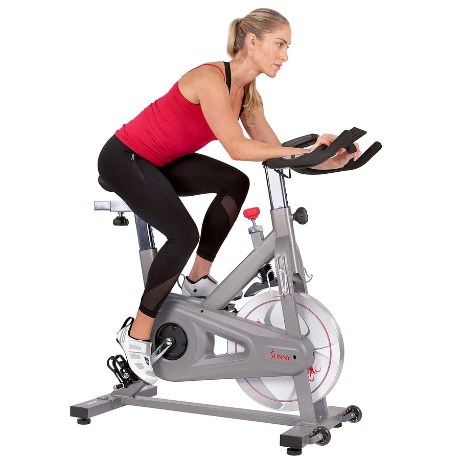 A woman in a red tank top and black leggings intensely rides the Sunny Health & Fitness Synergy Series Magnetic Indoor Cycling Exercise Bike, her eyes fixed forward as she grips the handlebars, showcasing her dedication beyond just exercise.