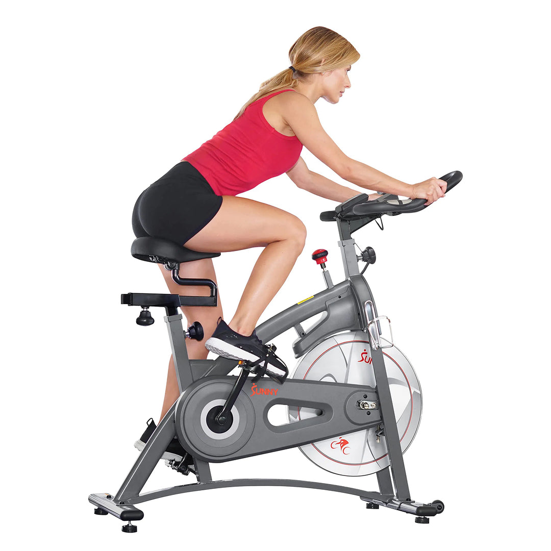 Wearing a red tank top and black shorts, a person vigorously pedals the Sunny Health & Fitness Synergy Series Magnetic Indoor Cycling Exercise Bike. The gray bike with red accents enhances the intensity of their focused workout session.