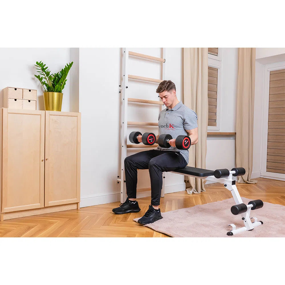 A man exercises on a BenchK Pro Gym Swedish Ladder 723 in his home gym, lifting adjustable dumbbells. Wearing a gray T-shirt and black pants, he focuses intently. In the background are a wooden cabinet with a potted plant and the wall-mounted pull-up bar from the set.