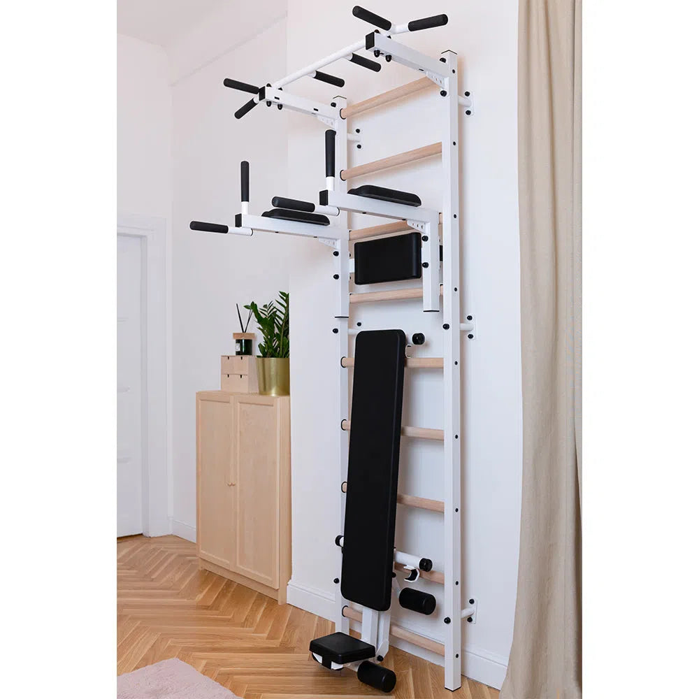 The BenchK Pro Gym Swedish Ladder (723) includes a pull-up bar, dip bar, and foldable bench. Its wall-mounted in a home setting next to a wooden cabinet and beige curtains for style and convenience.