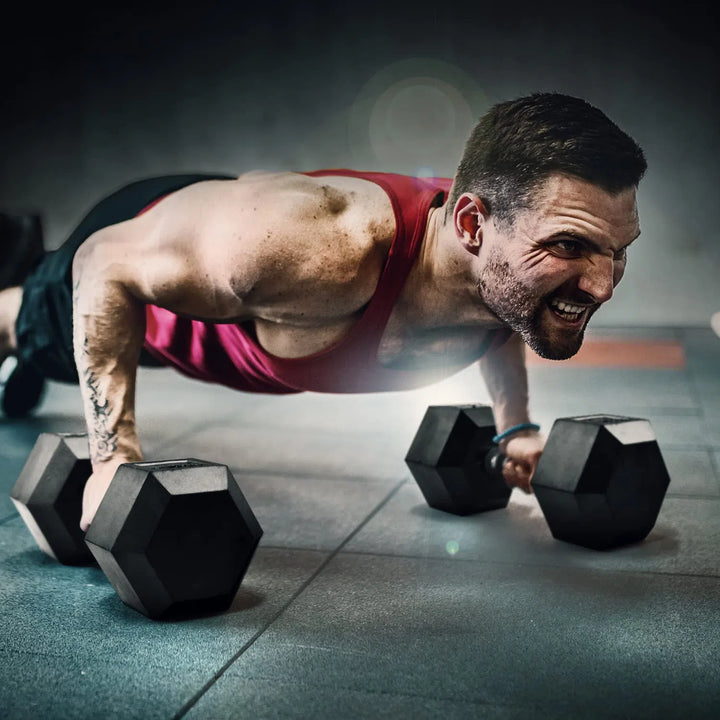 A man in a red tank top performs push-ups with Yes4All Rubber Grip Encased Hex Dumbbells on the gym floor. His focused expression and muscular form are highlighted, showcasing the premium rubber-coated, solid cast-iron core weights by Yes4All against a blurred background.