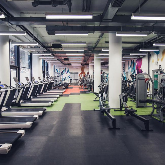 A spacious gym with rows of treadmills on the left, and various strength training machines on the right. The floor is covered with rubber mats in different colors. Large windows line the left wall, and there is graffiti on the back wall.